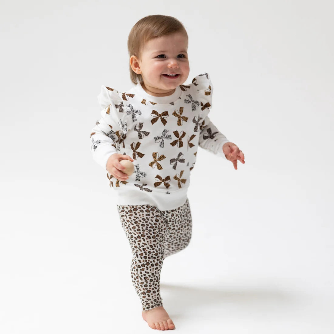 A smiling toddler walking barefoot on a white studio background, wearing a white long-sleeve top with brown bow prints and Angel Dear brown leopard-print leggings.