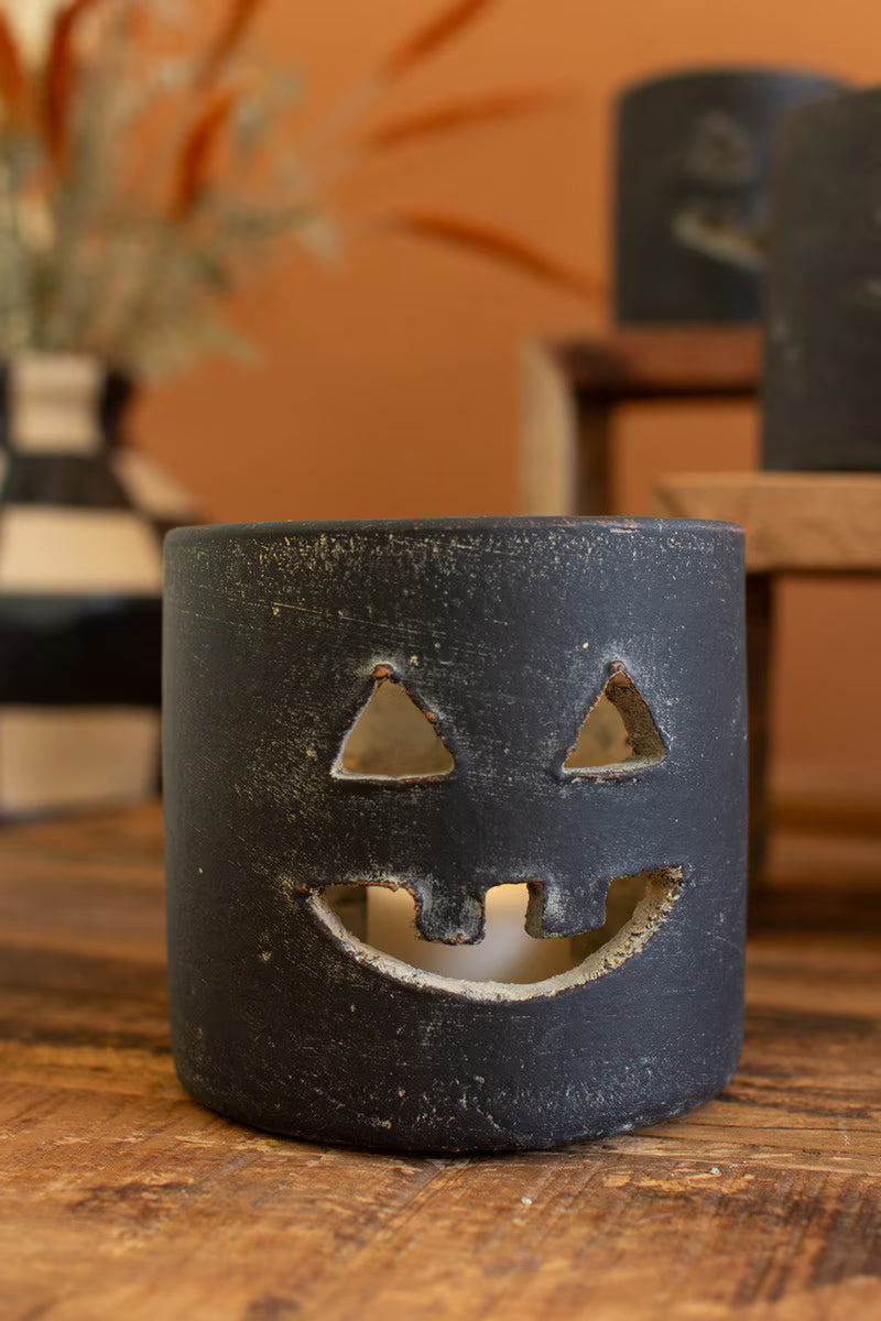 A black clay jack-o'-lantern candle holder with a textured finish, featuring cutouts for a face, sitting on a wooden table with a warm orange-brown background.