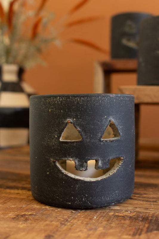 A black clay jack-o'-lantern candle holder with a textured finish, featuring cutouts for a face, sitting on a wooden table with a warm orange-brown background.