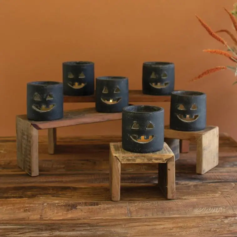 Six black clay candle holders with carved jack-o'-lantern faces, displayed on rustic wooden risers against a warm orange-brown background.