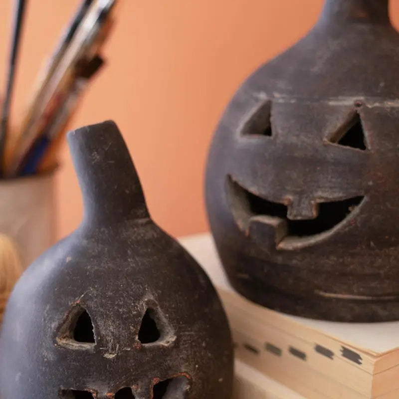 Two dark carved jack-o'-lantern-shaped ceramic vessels with cutout faces, displayed on stacked books against a warm peach background.
