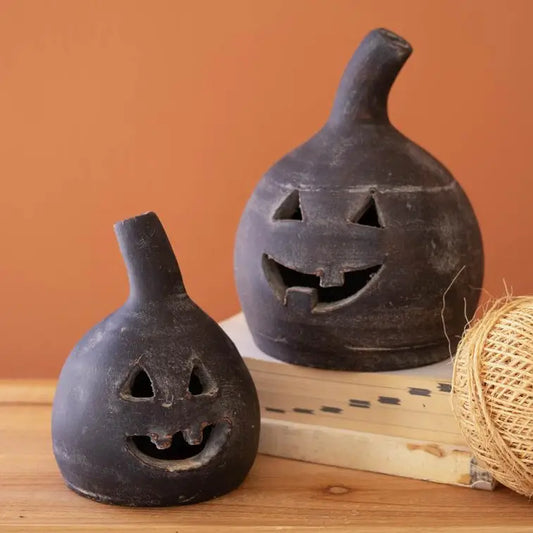 Two black clay pumpkin-shaped candle holders with carved jack-o'-lantern faces, styled on a wooden surface with a book and twine against a solid terracotta background.