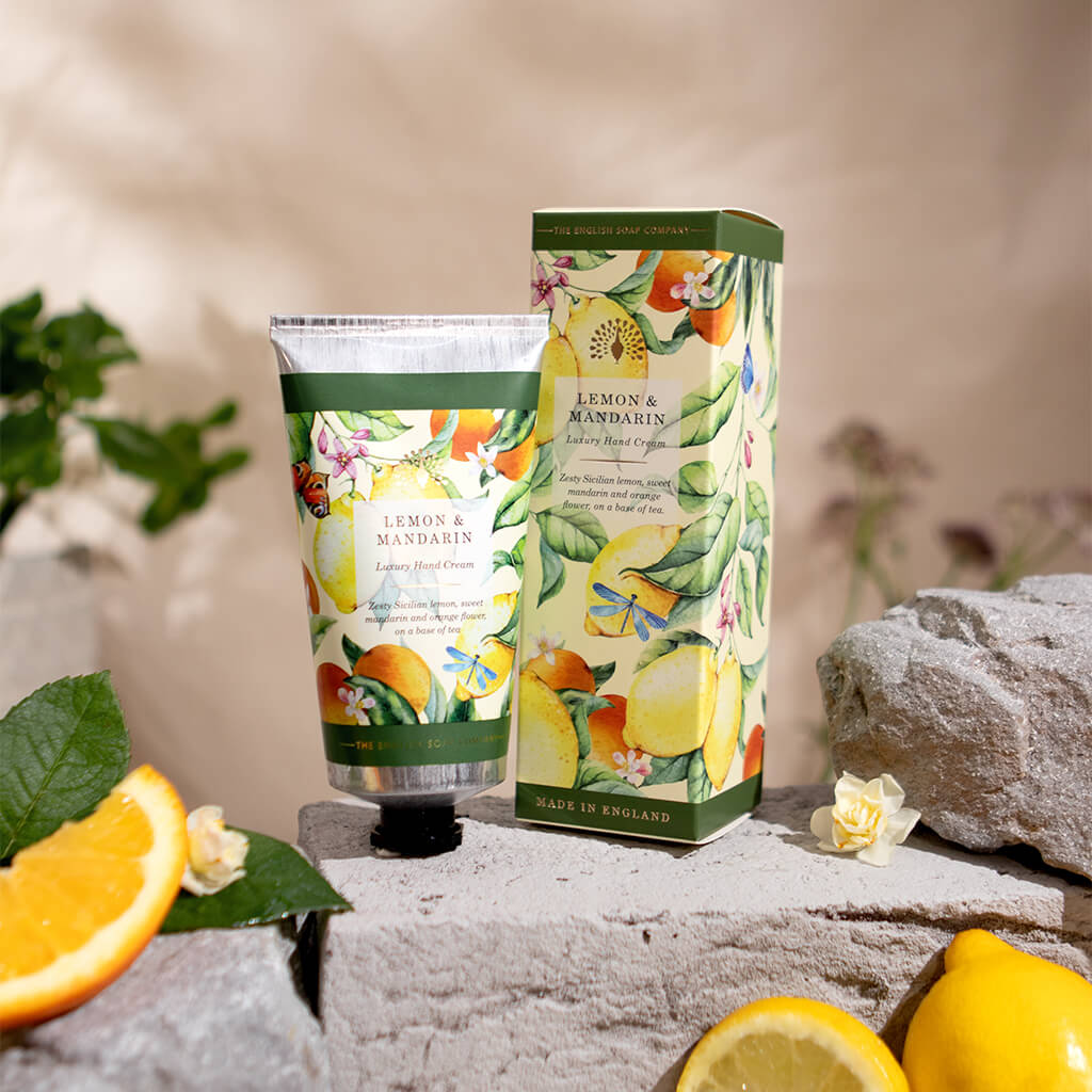 A tube and matching box of Lemon & Mandarin Hand Cream from The Handmade Soap Company, styled on a stone surface with lemons, oranges, and green leaves.
