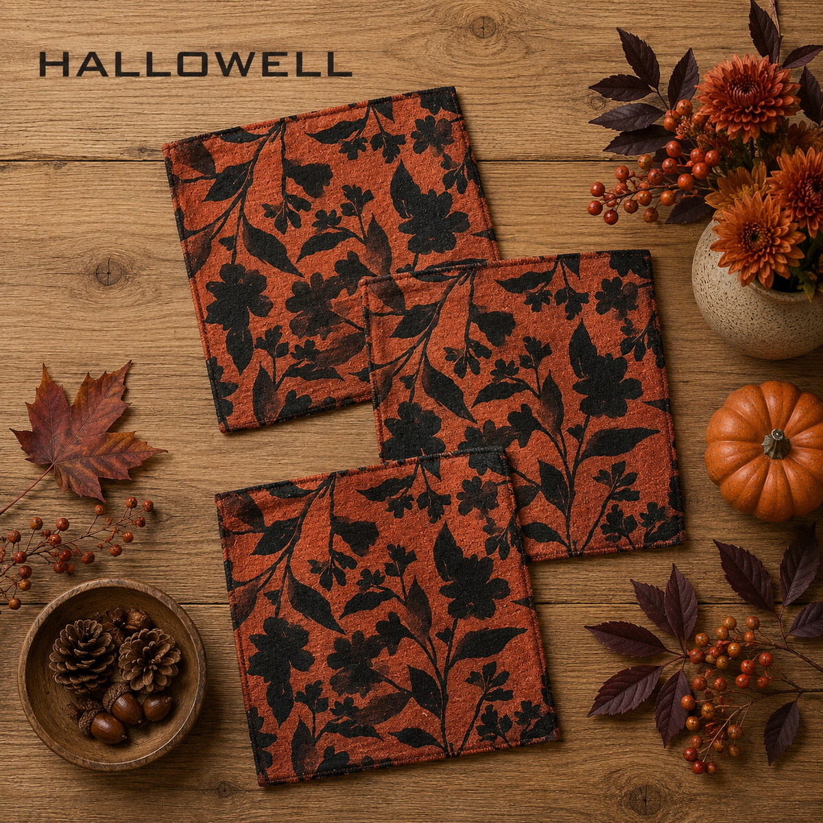 Geometry Hallowell dishcloth set of Three, square  with a black floral vine pattern on a rust-orange background, arranged on a wooden tabletop with autumn props including leaves, berries, pinecones, and a small pumpkin. The word HALLOWELL appears at the top left.  The little Exchange SKU 150069