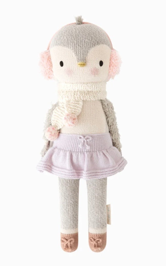 A plush penguin doll wearing pink pom-poms, a cream scarf, a light pink tutu skirt, and gray socks with pink shoes, photographed against a plain white background.