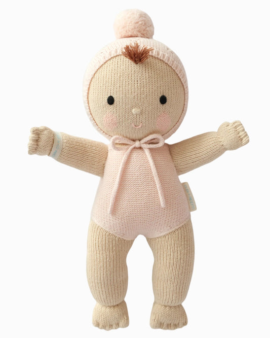 A knitted baby doll with a pale pink outfit, cream-colored limbs, a matching knit bonnet with pom-pom, and small black facial features, shown on a plain white background.