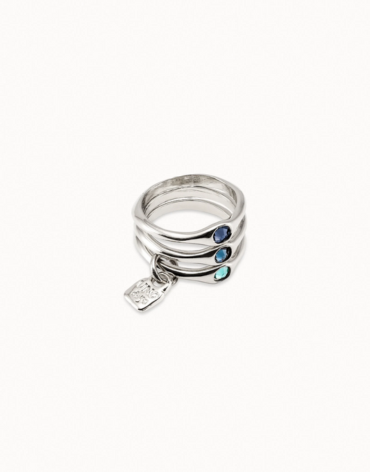 A triple sterling silver-plated ring with three blue Swarovski crystals, accompanied by a small silver tag with the brand 'UNO de 50' on it.