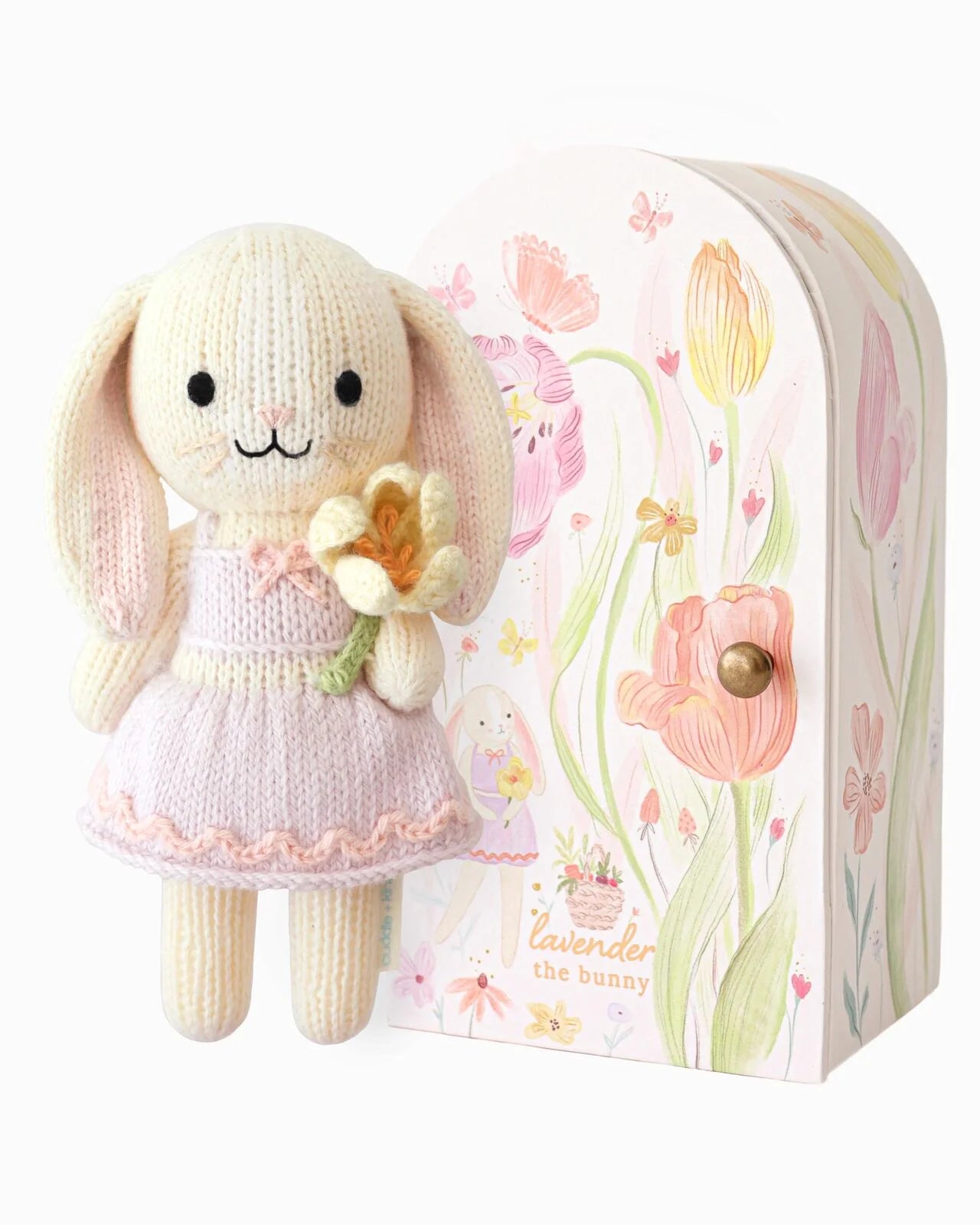 Knitted bunny toy with a pink dress holding a flower, standing next to a decorative box with floral designs.