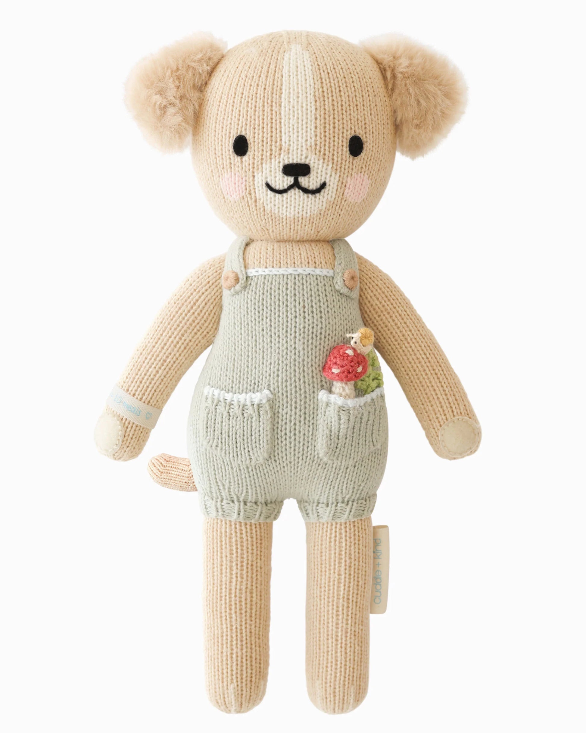 Knitted tan puppy wearing green overalls with a mushroom with a snail in his pocket