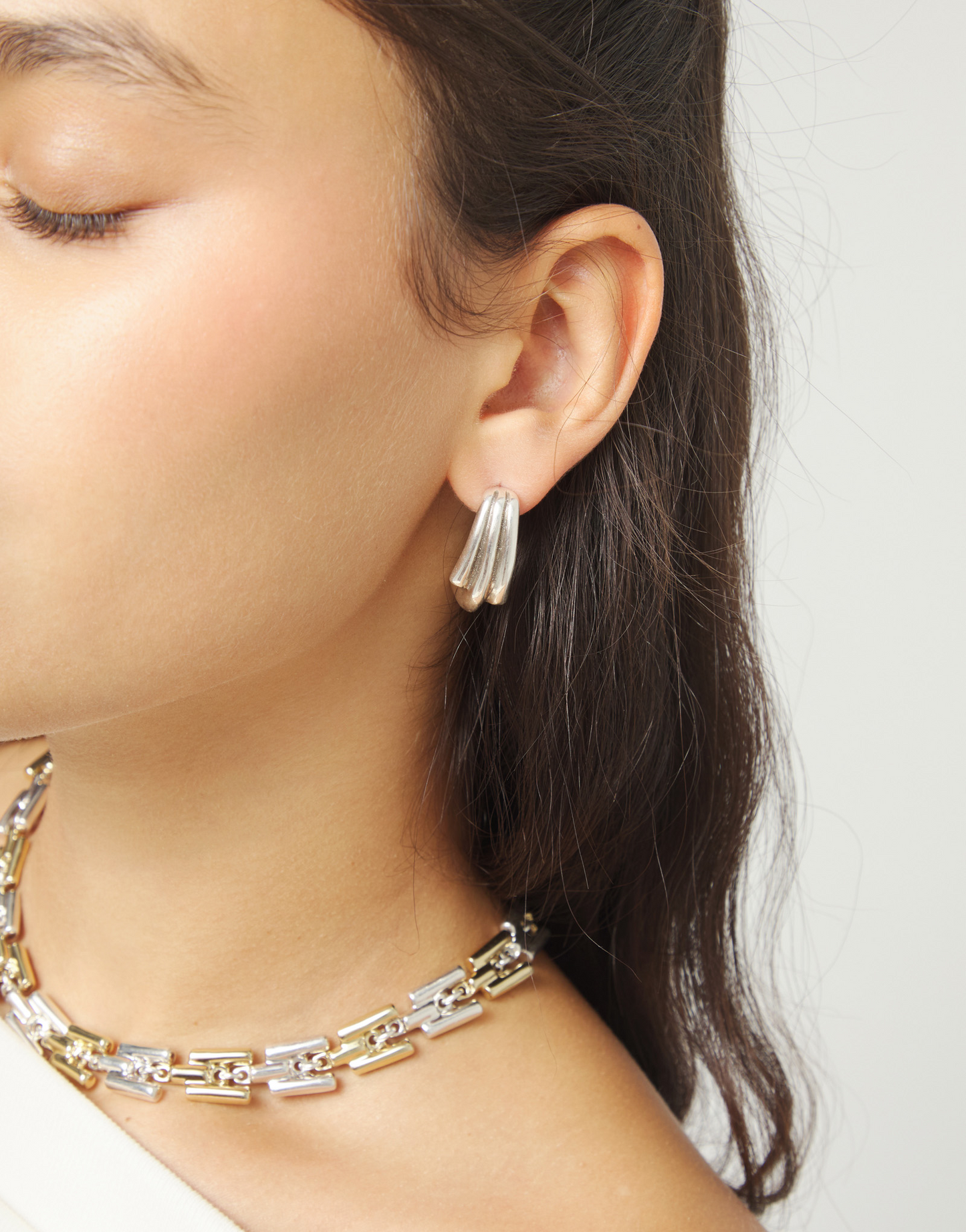 Close-up of a person wearing roots silver drop UNO de 50 earrings and a gold and silver necklace on a plain background