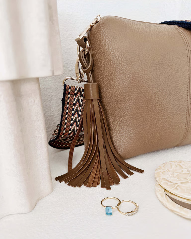 A close-up of a Louenhide Kasey Crossbody Bag latte pebbled leather shoulder bag with a long brown leather tassel charm, styled on a white textured surface with rings and a small woven pouch.