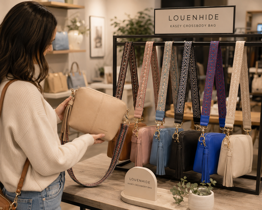 A woman in a cream ribbed sweater and blue jeans holds a beige crossbody bag while standing in a boutique. A black display rack behind her shows multiple color and pattern variations of the Louenhide Kasey Crossbody Bag, including pink, blue, black, and purple straps with tassels.