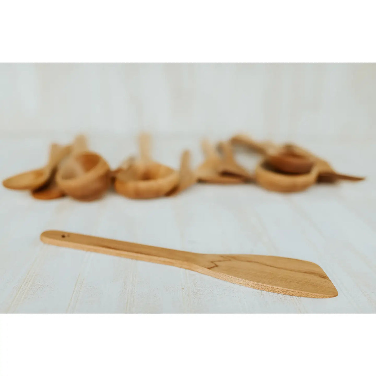 A maple light natural wood spatula with a small hole in the handle by holland bowl mill, shown in sharp focus in the foreground with a blurred row of similar wooden utensils in the background on a white surface.
