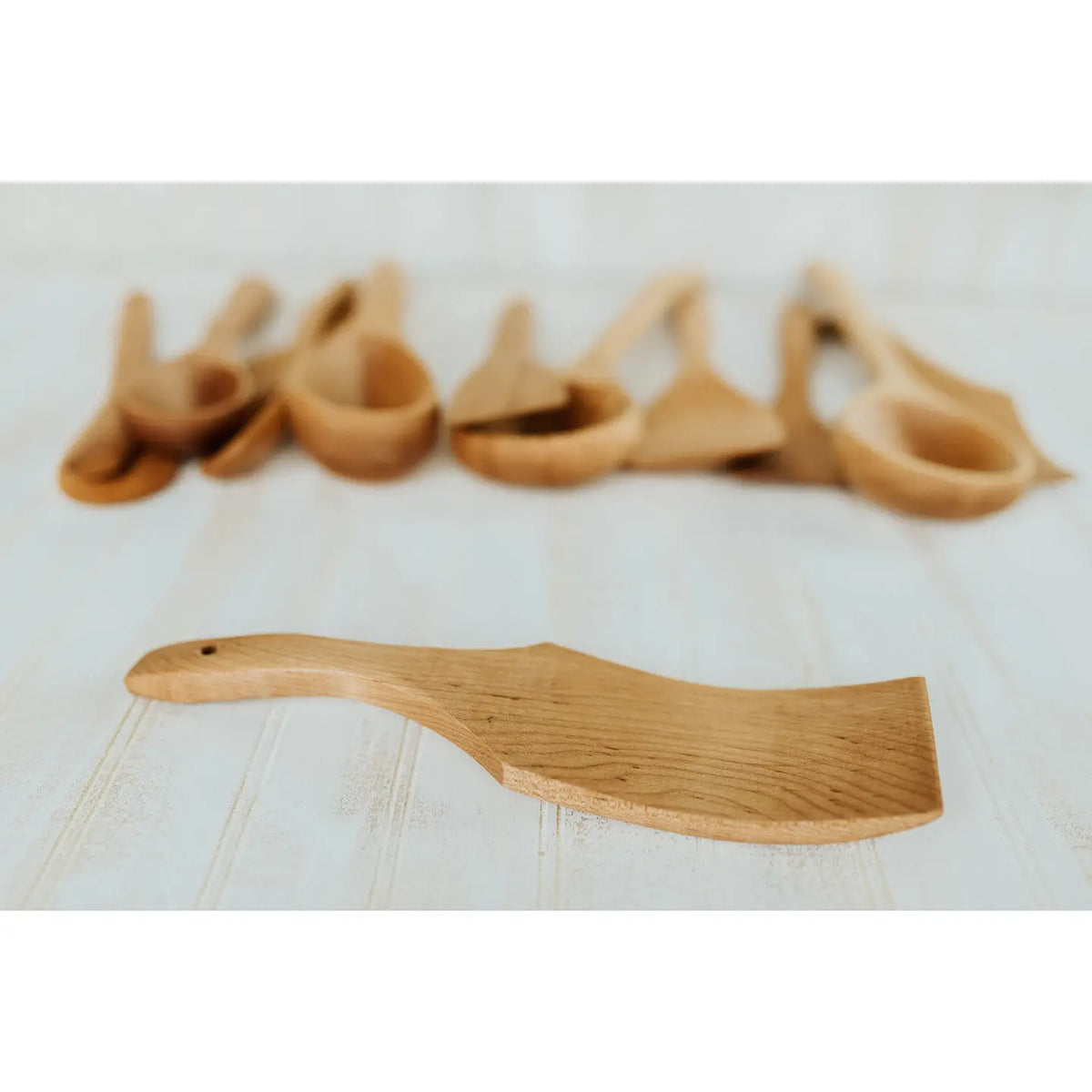 A light natural wood serving spatula with a wide flat blade and handle, shown in sharp focus in the foreground with several similar wooden utensils blurred in the background on a white surface.