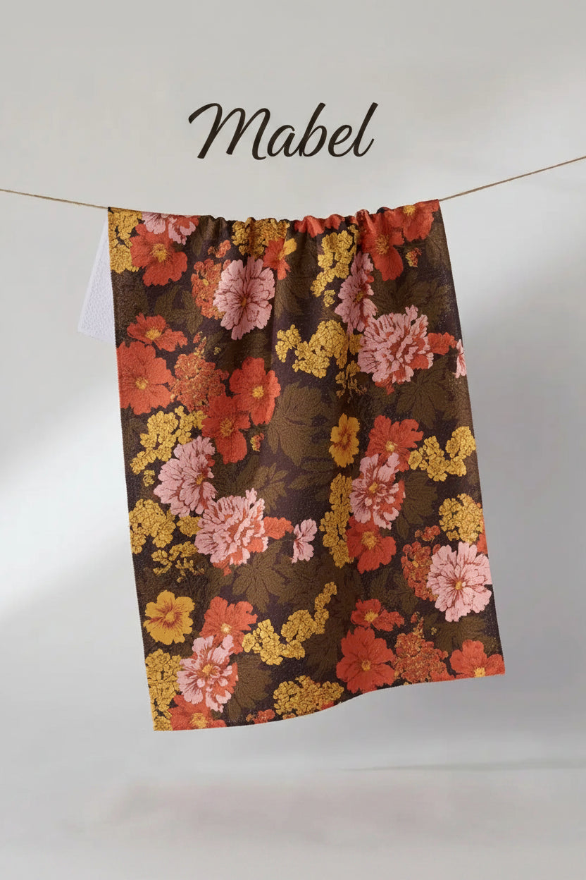 A Geometry Mabel tea towel with a vintage floral pattern in orange, pink, and yellow on a dark brown background, hanging from a thin string against a light gray wall.