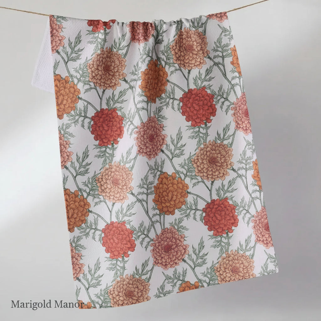A white kitchen Geometry Marigold manor tea towel with a repeating floral print of orange, red, and peach marigold flowers with green leaves, hanging from a thin cord against a light gray background.