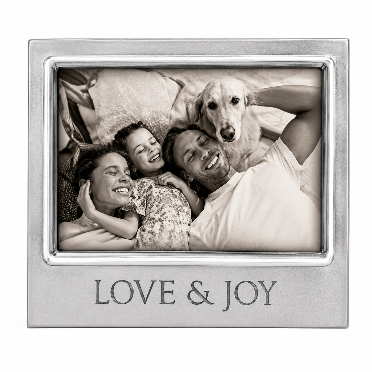 A Mariposa 4300LI Love & Joy Signature 4x6 silver-toned photo frame with a polished metal border and the words 'LOVE & JOY' engraved at the bottom. The frame holds a black-and-white photograph of a smiling family of four lying down with a golden retriever dog.