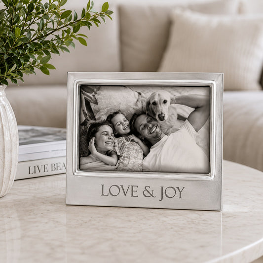 A Mariposa 4300LI Love & Joy Signature 4x6 rectangular silver picture frame with the words 'LOVE & JOY' engraved on the front, holding a black-and-white photo of a couple and their dog. The frame sits on a light marble table in a cozy living room setting next to a vase of greenery and books.