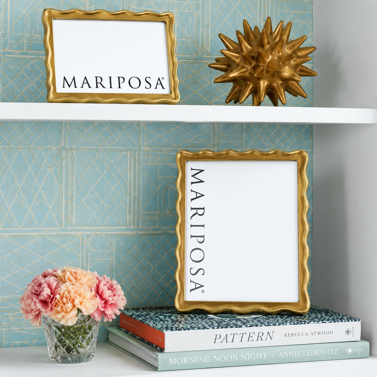 Two gold-framed picture frames with white mats and the word 'MARIPOSA' printed vertically or horizontally, styled on a shelf with a gold spiky decor object, books, and pink flowers against a blue patterned wall.