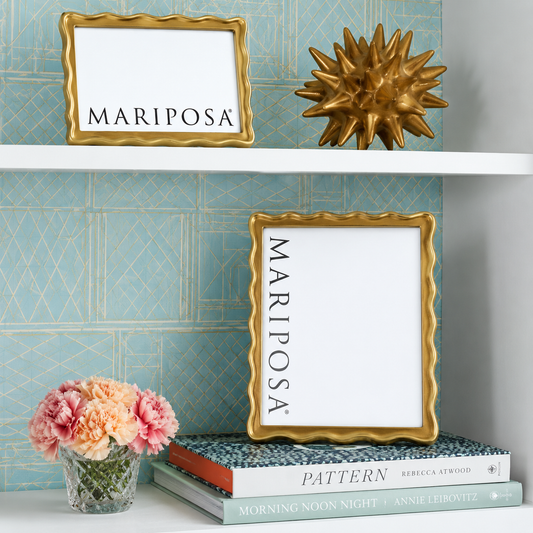 Two gold-framed picture frames with white mats and the word 'MARIPOSA' printed vertically or horizontally, styled on a shelf with a gold spiky decor object, books, and pink flowers against a blue patterned wall.