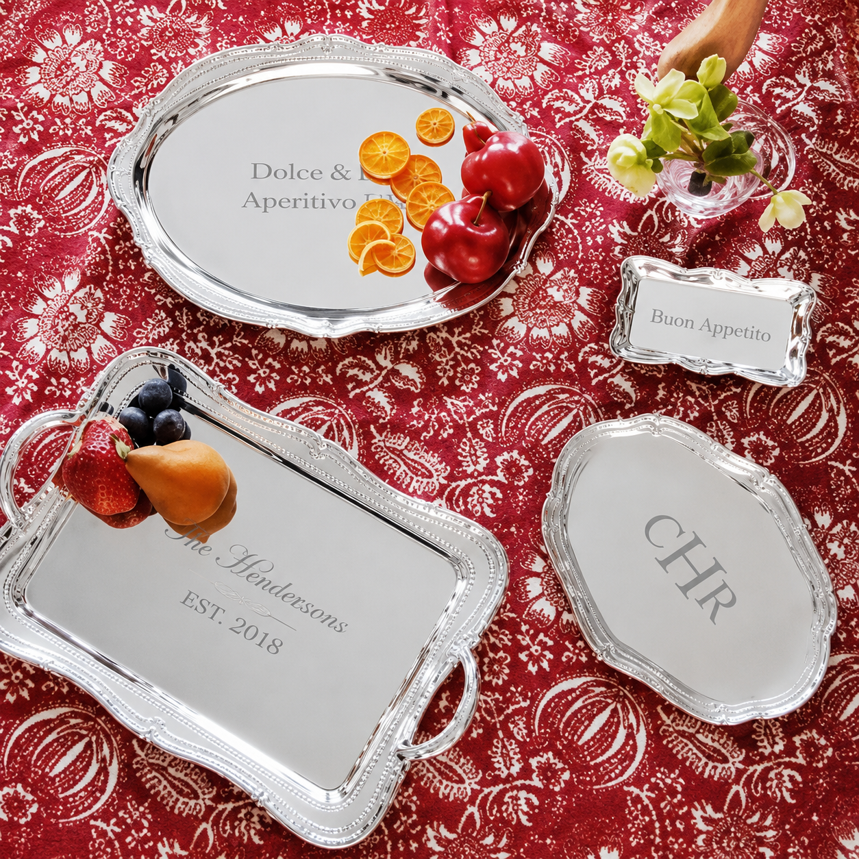 A set of personalized Mariposa Sueno silver serving trays arranged on a red patterned tablecloth, with fruit and flowers used as styling props. The trays include oval, rectangular, and scalloped shapes, some engraved with names like Dolce & Aperitivo, Chr.
