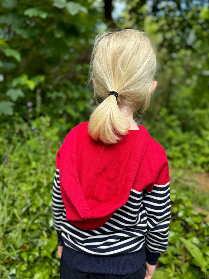 Toddler wearing a red hooded me and Henry jacket with a striped shirt underneath, standing in a green outdoor setting.