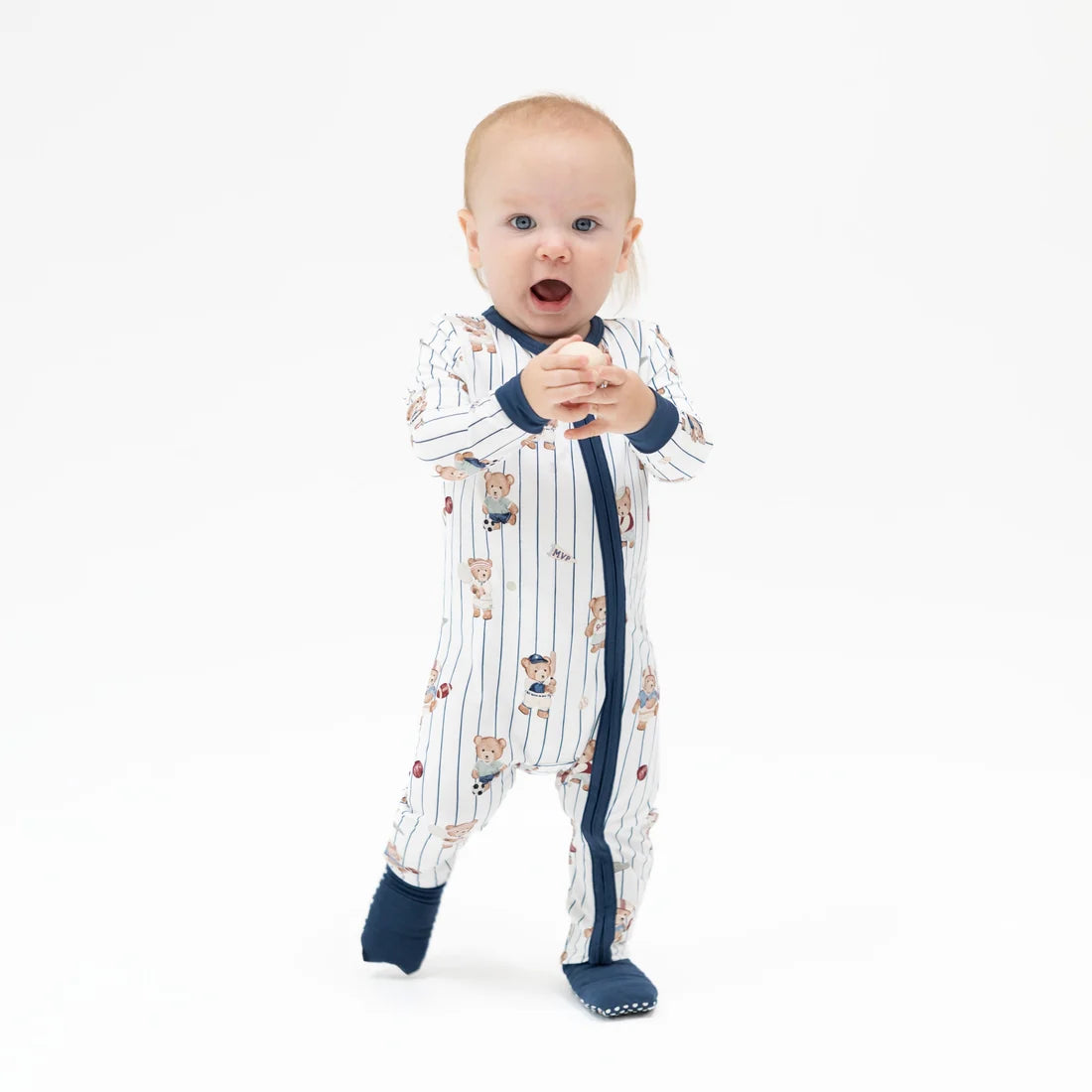 A baby standing on a white studio background wearing a white Angel dear mixed sports bear footless sleeper with navy blue trim and a teddy bear print, along with navy socks.