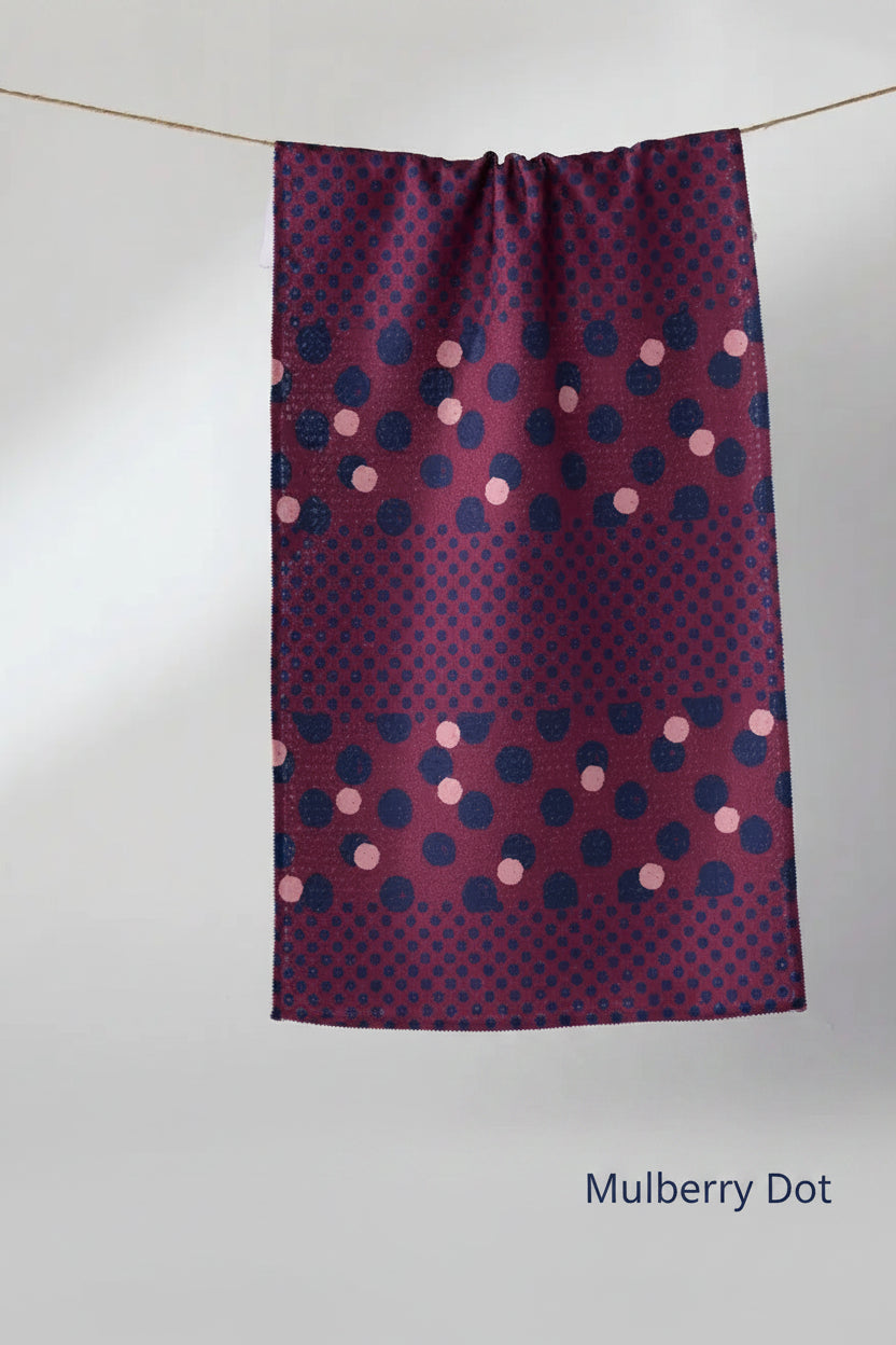 A rectangular kitchen Geometry mulberry dot tea towel with a dark purple and navy blue polka dot pattern and pink accents, hanging from a thin string against a light gray background.
