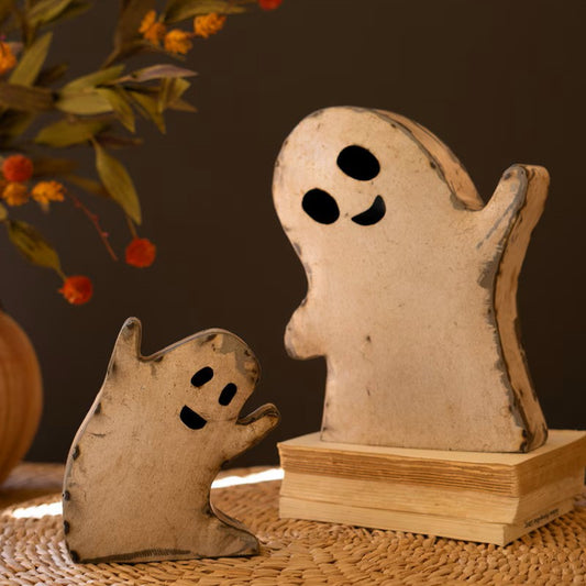 Two rustic ghost-shaped decorative metal sculptures in a weathered cream and brown finish, one standing on a small book pedestal and one smaller beside it, set on a woven surface against a dark brown background with blurred orange flowers.