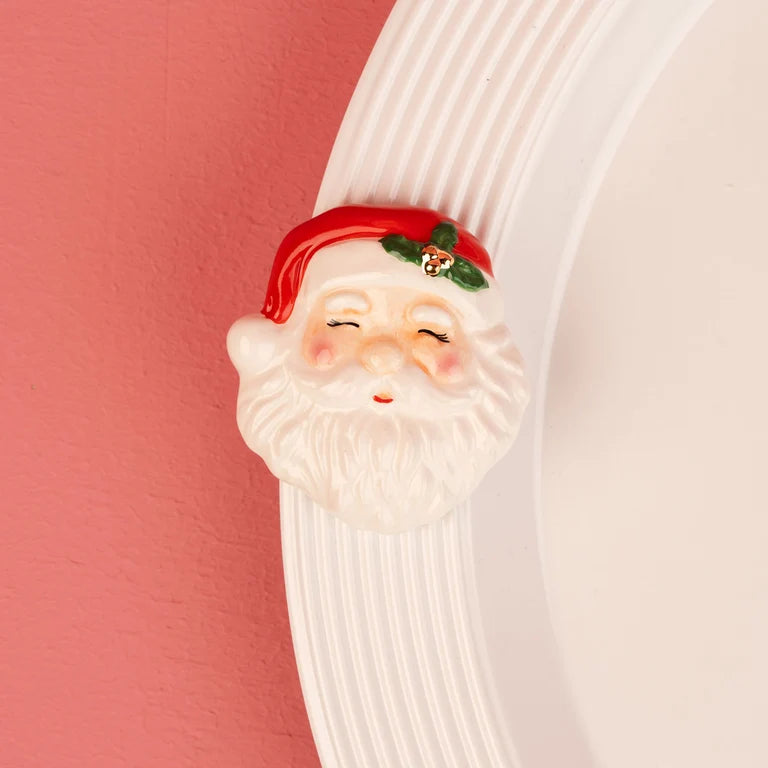 A Nora Fleming mini Santa Claus face with a red hat, white beard, and small green holly accent, shown mounted on a white ribbed circular plate