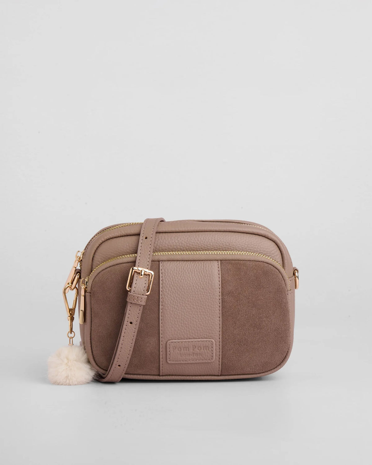 A taupe-brown crossbody bag with a dual-texture design featuring pebbled leather and suede panels, gold-tone hardware, a matching shoulder strap, and a fluffy pom-pom charm, set against a plain light gray background.