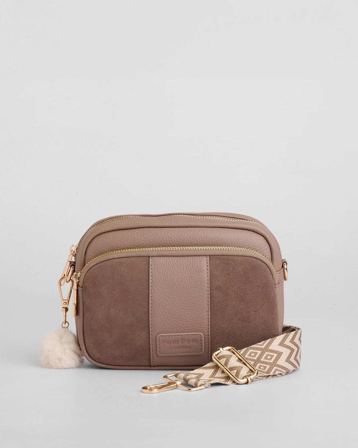 A taupe brown crossbody bag with gold-tone hardware, leather and suede panel design, a fluffy pom-pom charm, and a patterned woven strap on a light gray background.
