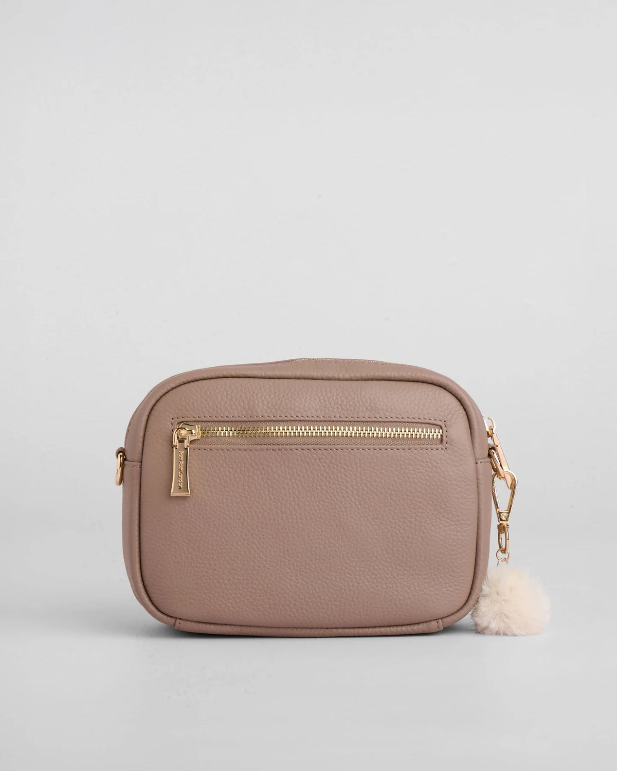 A small rectangular crossbody bag in textured mocha leather with gold-tone hardware, a front zip pocket, and a fluffy pom-pom charm, shown on a light gray background.