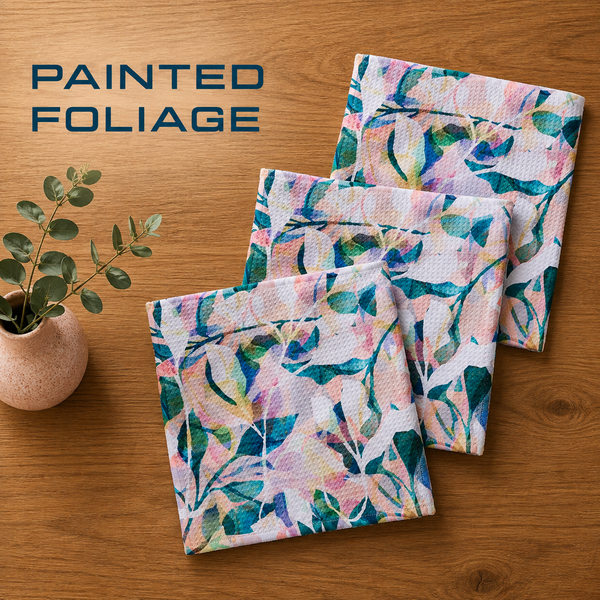 Geometry painted foliage dishcloth set with Three square fabric pockets with a painted watercolor foliage pattern in shades of teal, green, pink, and blue on a white background, arranged on a wooden table next to a small potted plant. The text 'PAINTED FOLIAGE' is visible in the top left corner.  The Little Exchange SKU 141356