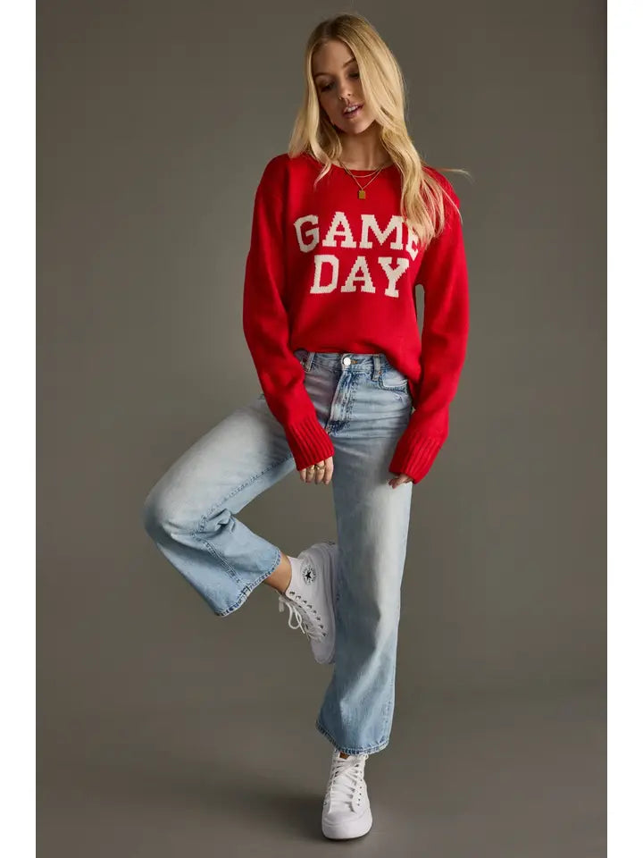 Woman wearing a red 'GAME DAY' Panache Sweater with blue jeans and white sneakers on a gray background