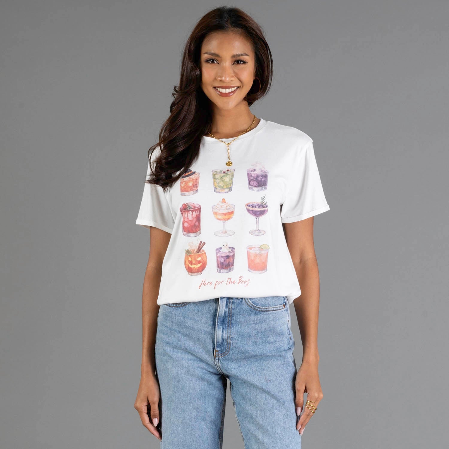 A woman modeling a white short-sleeve graphic t-shirt with illustrations of colorful Halloween drinks and the text 'here for the boos', paired with light blue jeans against a solid gray studio background.