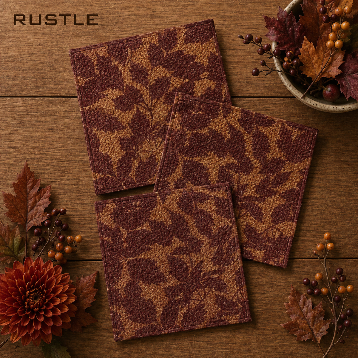 Geometry set of Three square Rustle dishcloths with a brown and tan autumn leaf pattern, arranged on a wooden tabletop with fall leaves and berries.  The Little Exchange SKU 150102