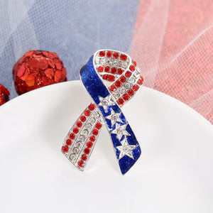 A patriotic awareness ribbon brooche pin featuring red, white, and blue rhinestone detailing and silver-tone metal, displayed on a white plate with festive red ornaments in the background.