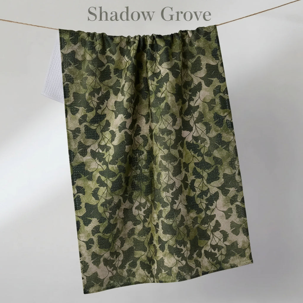 A rectangular Geometry shadow grove tea towel with an all-over ivy leaf pattern in shades of green and beige, hanging from a thin cord against a plain light gray background.