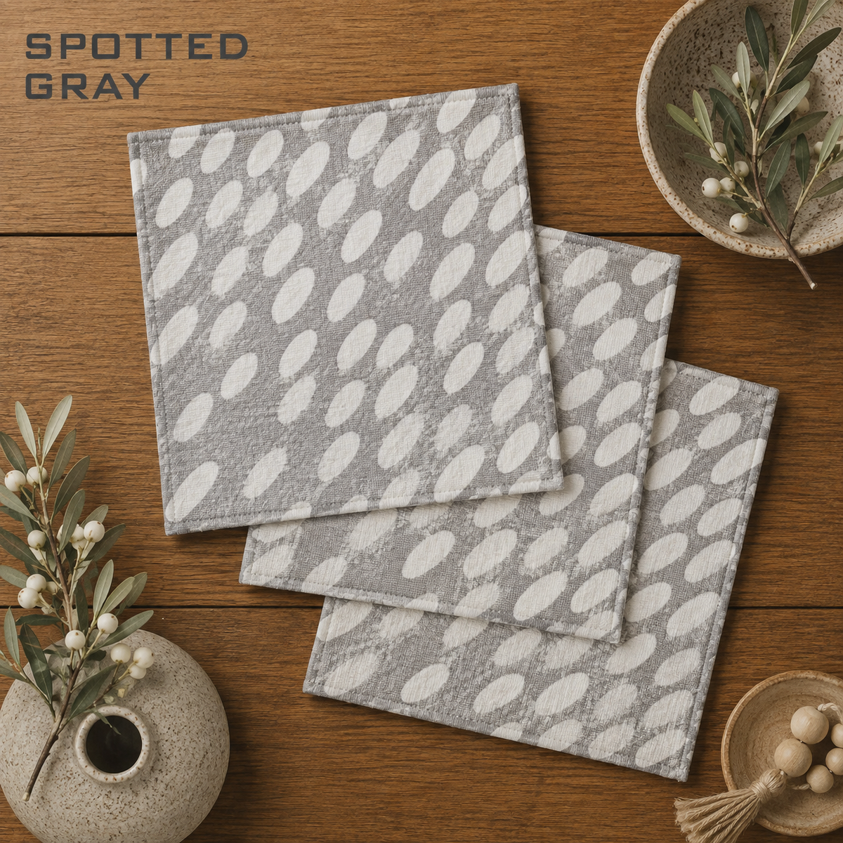 Geometry Spotted Gray square dishcloth set of three with a white spotted leaf pattern are arranged on a wooden tabletop with decorative vases and branches. The text 'SPOTTED GRAY' appears in the top left corner.  The Little Exchange SKU 14445