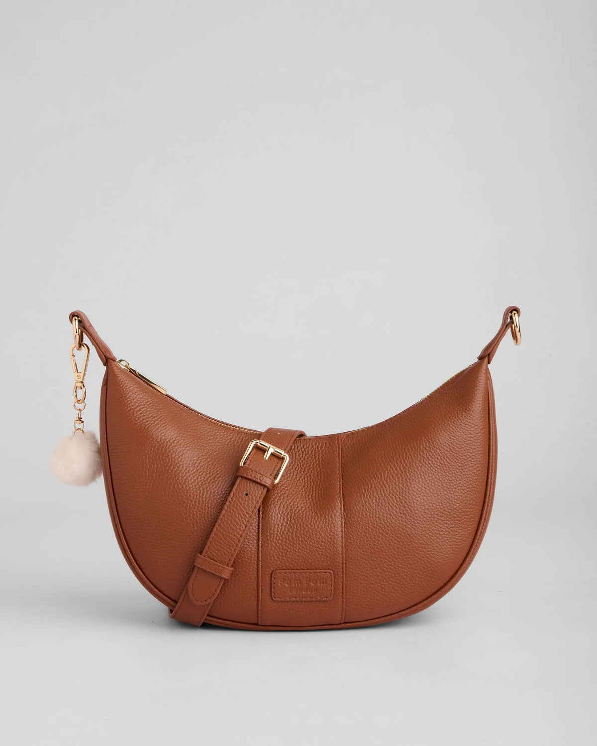 Brown chestnut leather handbag with a gold buckle and a pom pom tassel on a light gray background