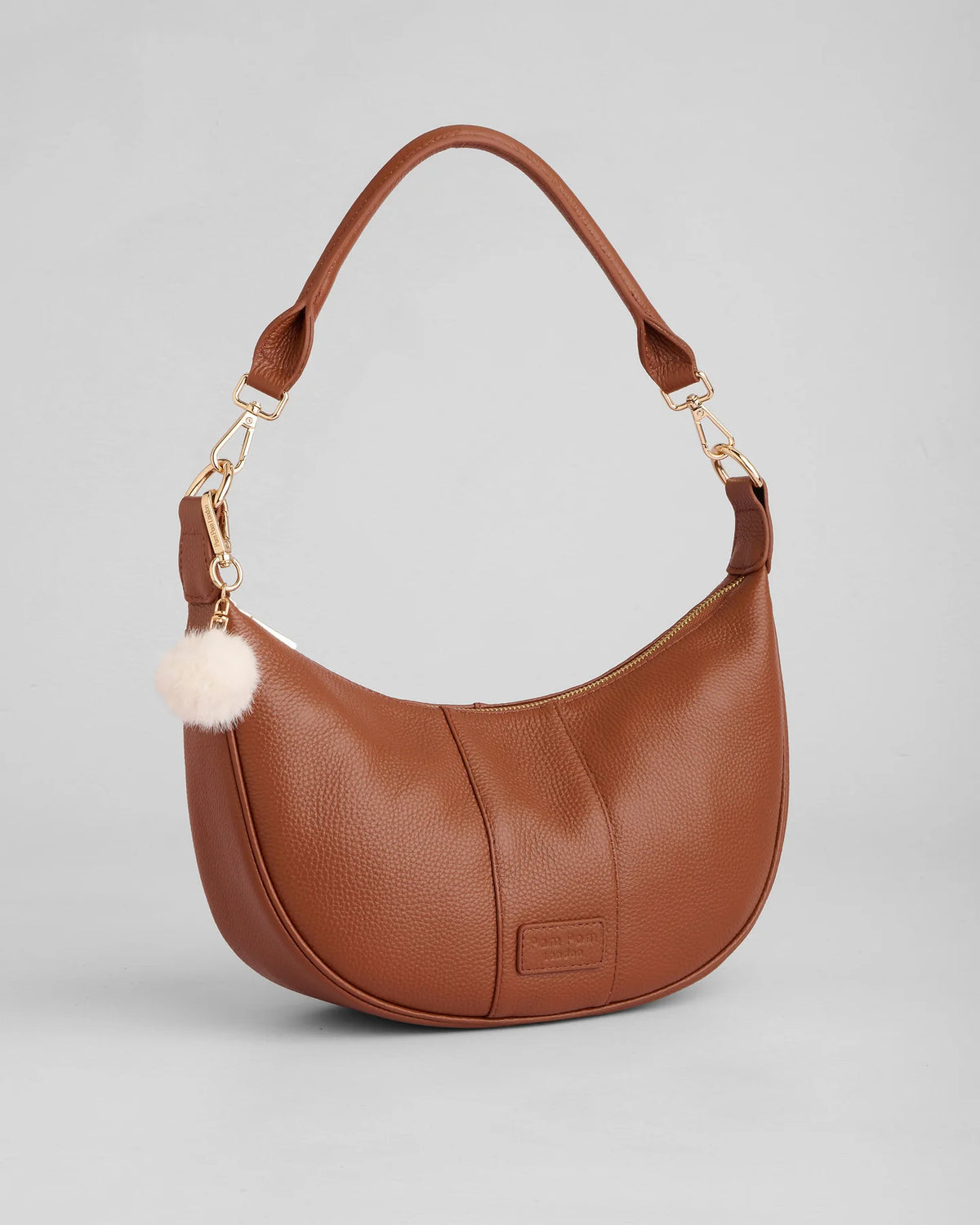 A brown pebbled leather crescent-shaped shoulder bag with a gold-tone clasp, a matching leather top handle, and a fluffy cream pom-pom charm, shown on a light gray background.