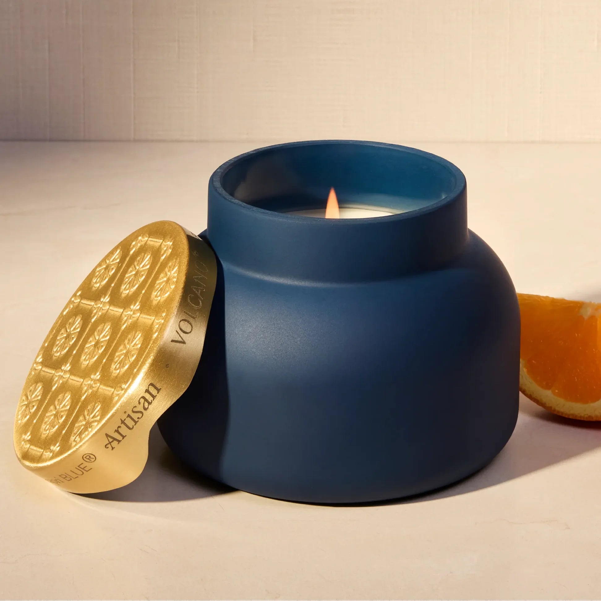 A matte navy blue jar candle with a gold lid leaning beside it on a light beige surface. A lit candle flame is visible inside the jar, and an orange slice sits to the right.