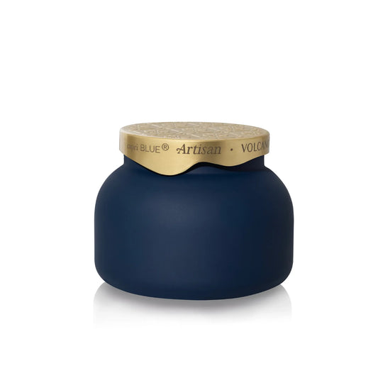 A dark navy blue jar with a textured gold lid, featuring the brand name 'Capri  Blue' and product line 'Artisan Volcano' printed on the gold band.