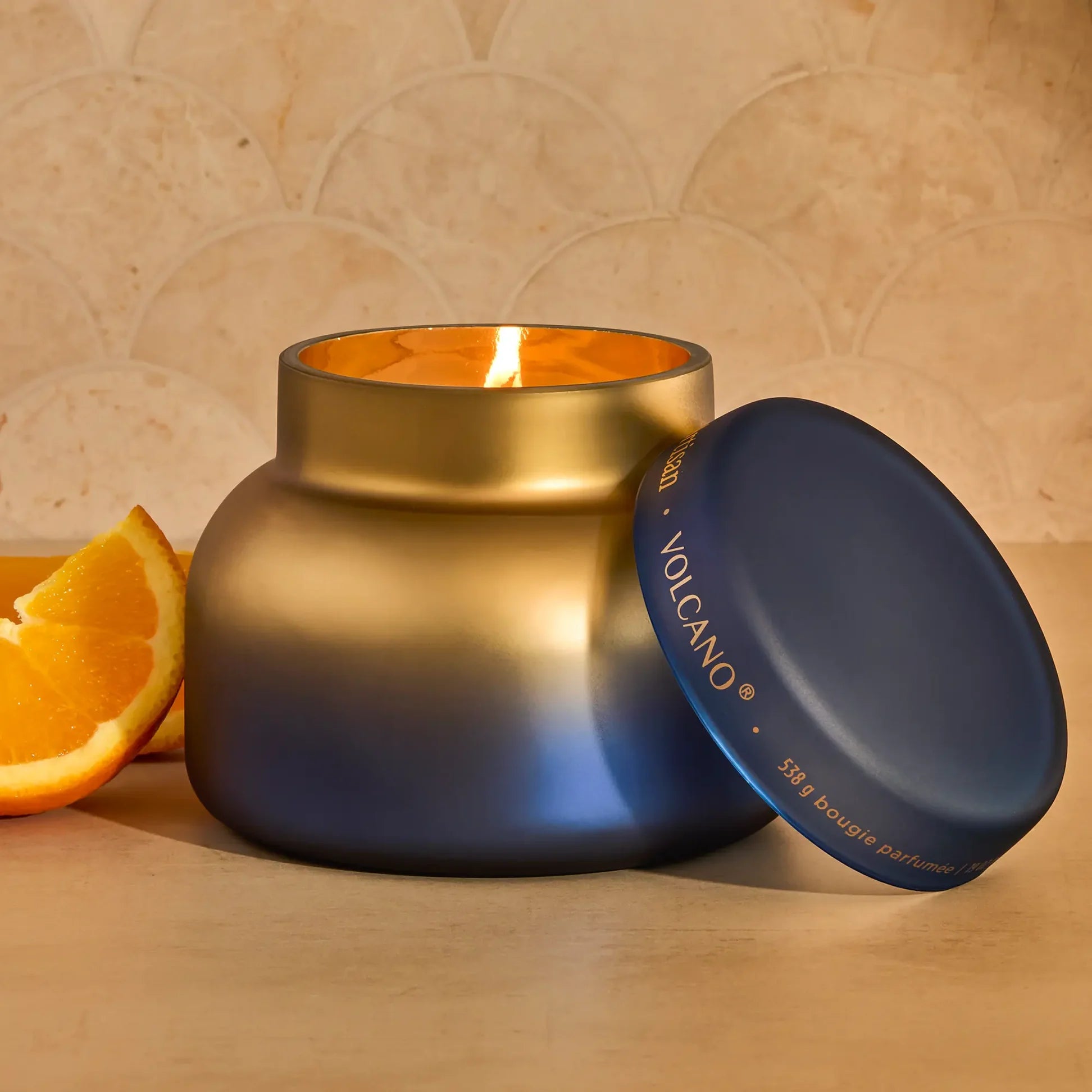A lit scented candle in a matte gradient jar that transitions from gold at the top to deep blue at the bottom, with its matching lid leaning against it. The jar sits on a light wooden surface next to a slice of orange, with a beige tiled wall in the background.
