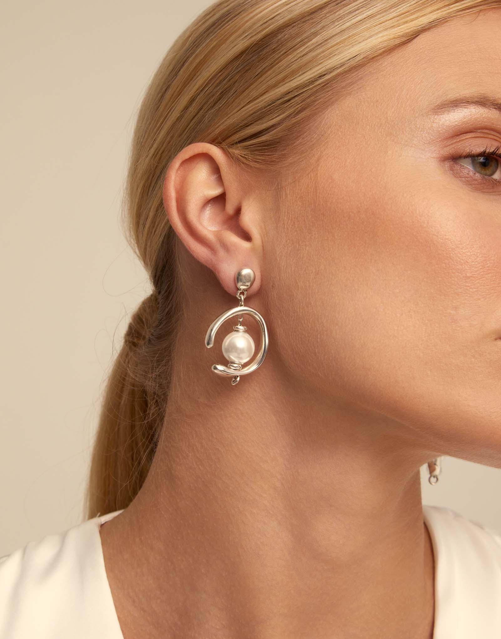 Close-up of a woman wearing UNOde50 Inorbit Silver Drop Earrings with Pearls on a beige background