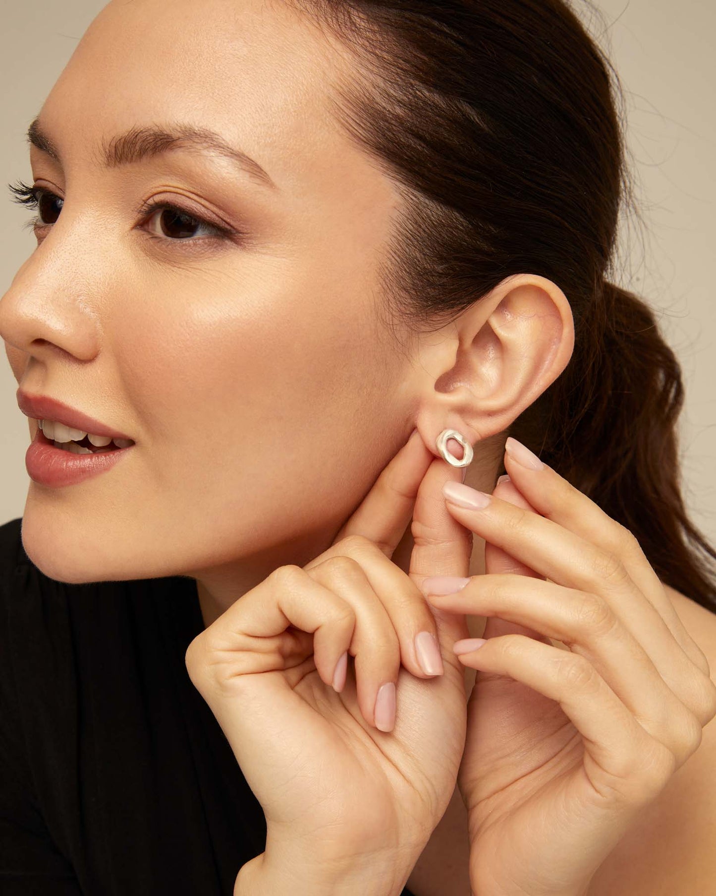 Woman adjusting an UNOde50 silver oval earring on her ear with a neutral background