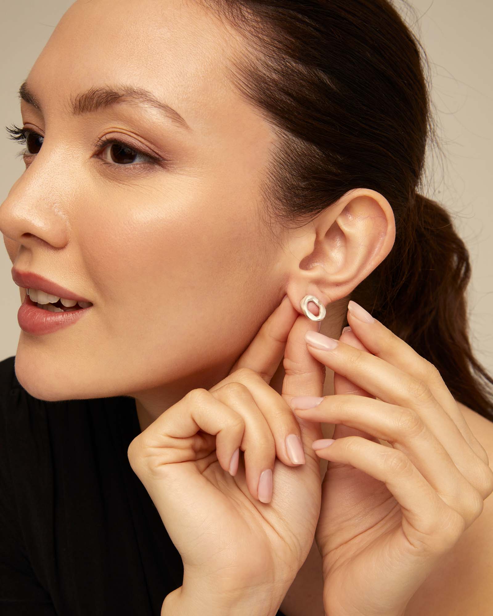 Woman adjusting an UNOde50 silver oval earring on her ear with a neutral background