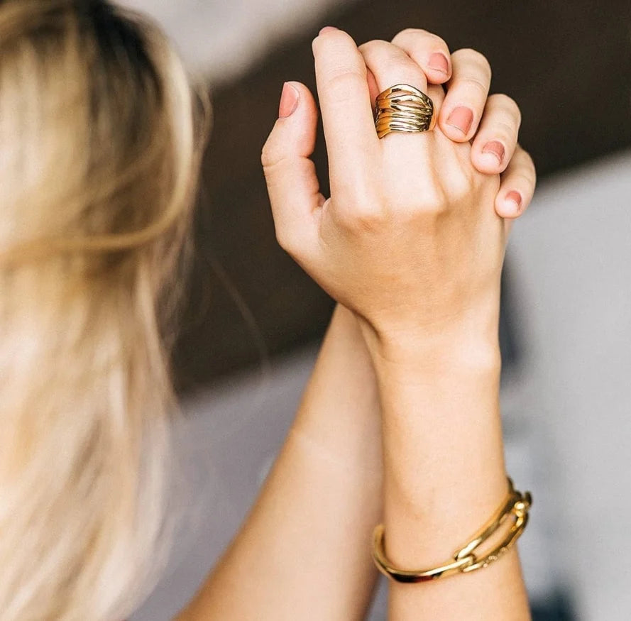 Close-up of two hands holding each other, wearing an UNOde50 Nihiwatu Beach Gold Stacked Ring and a bracelet.