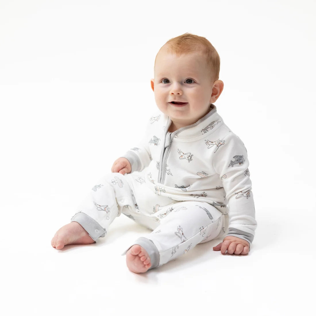A baby sitting on a white studio background wearing a white longangel dear vintage pilots long-sleeve footless sleeper with a gray zipper and a subtle gray animal print.